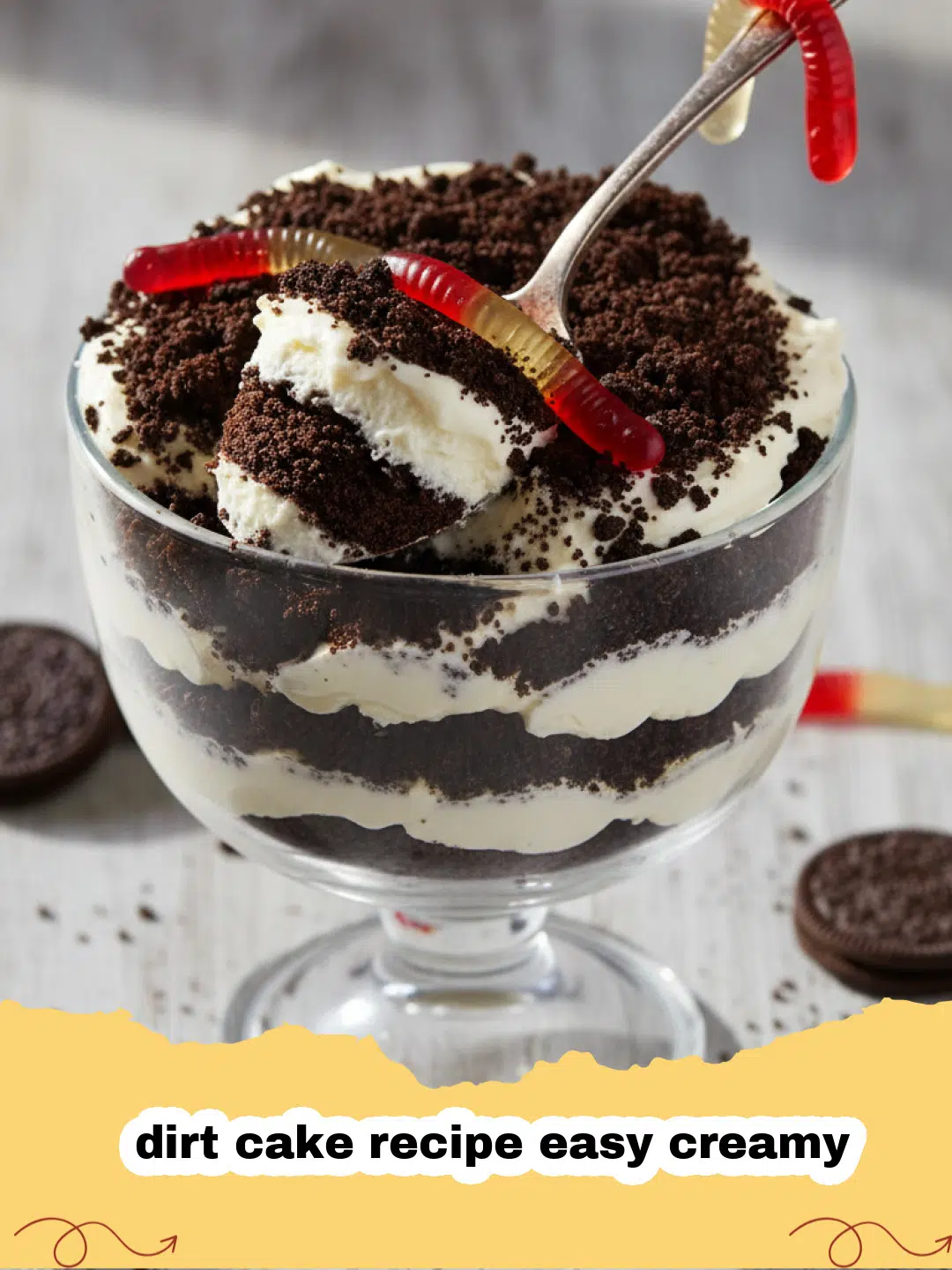 dirt cake recipe easy creamy - A clear glass trifle dish filled with layers of creamy dirt cake, topped with crushed Oreos and gummy worms.